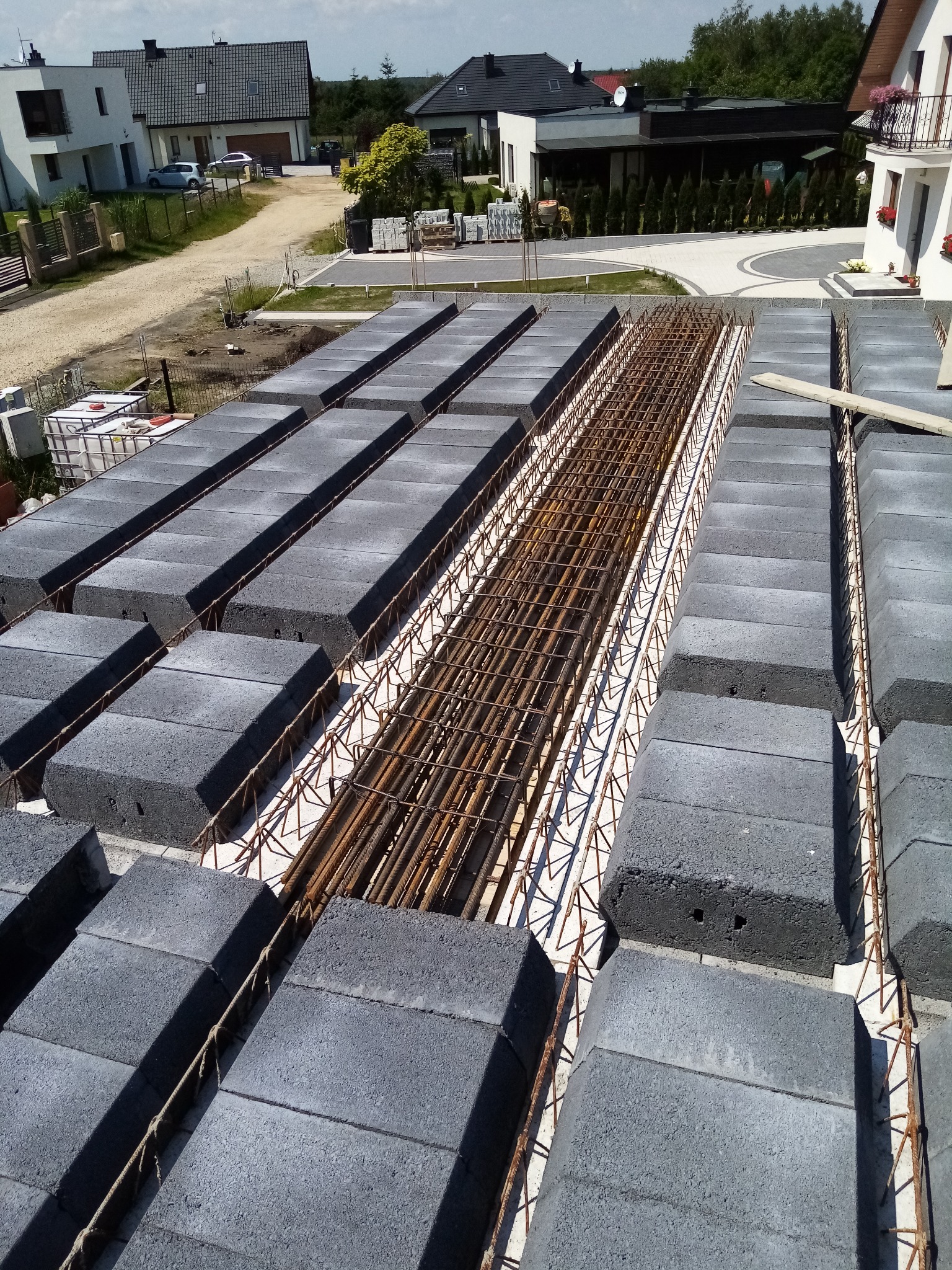 Wieniec stropowy w trakcie budowy, zbrojenie stalowe w szalunku z betonowych bloczków, widok z góry na budowę domu jednorodzinnego w słoneczny dzień.