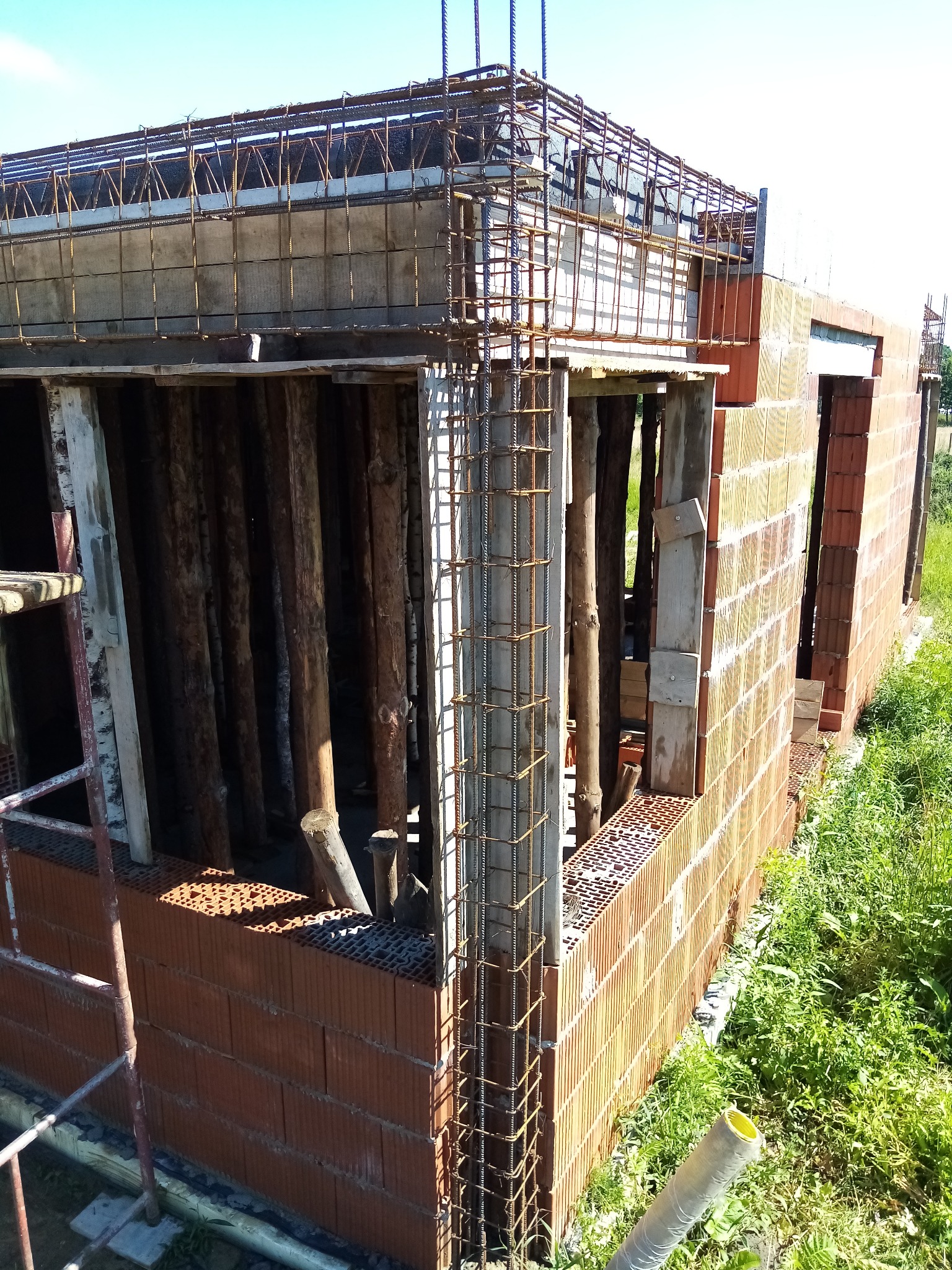 Budowa domu z cegły: ściany wzniesione, zbrojenie pod wieniec, drewniane stemple podtrzymujące strop, widok z zewnątrz.