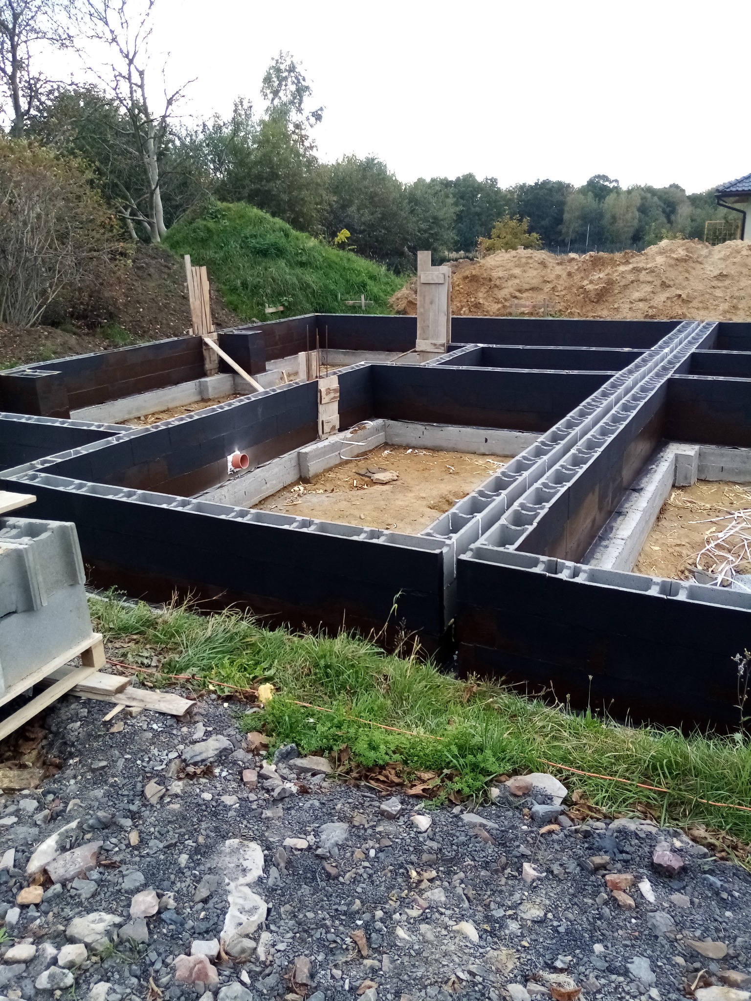Wykopane fundamenty nowego domu z bloczków betonowych, zabezpieczone czarną izolacją, widoczne zbrojenia i instalacje, w tle ziemia i zieleń.