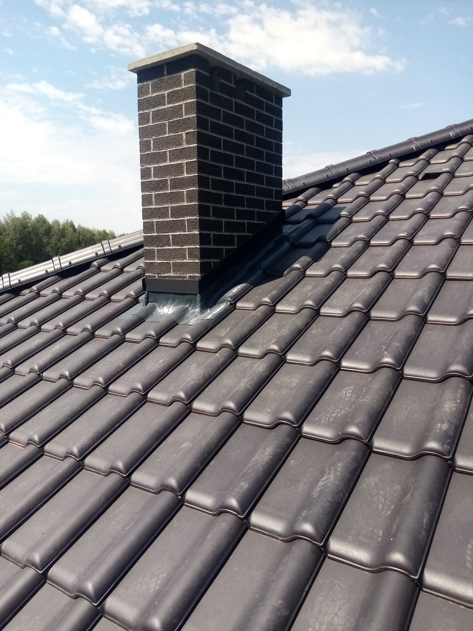 Ciemna dachówka ceramiczna z kominem z cegły klinkierowej na tle błękitnego nieba z chmurami.