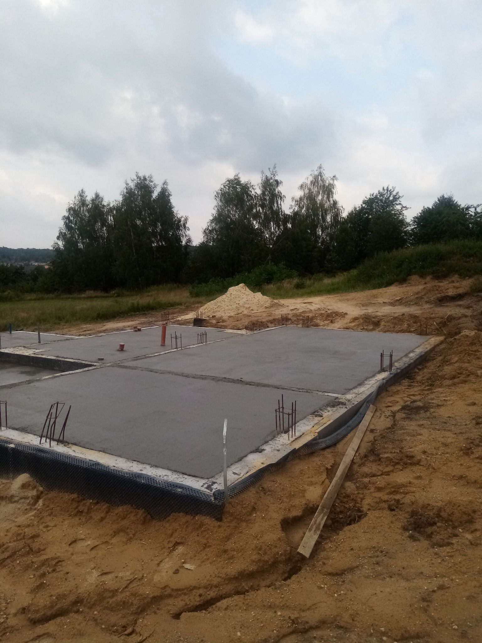 Zalany, świeży fundament płyty domu jednorodzinnego z wystającymi prętami zbrojeniowymi, zabezpieczony folią kubełkową, w tle drzewa i hałda piasku.