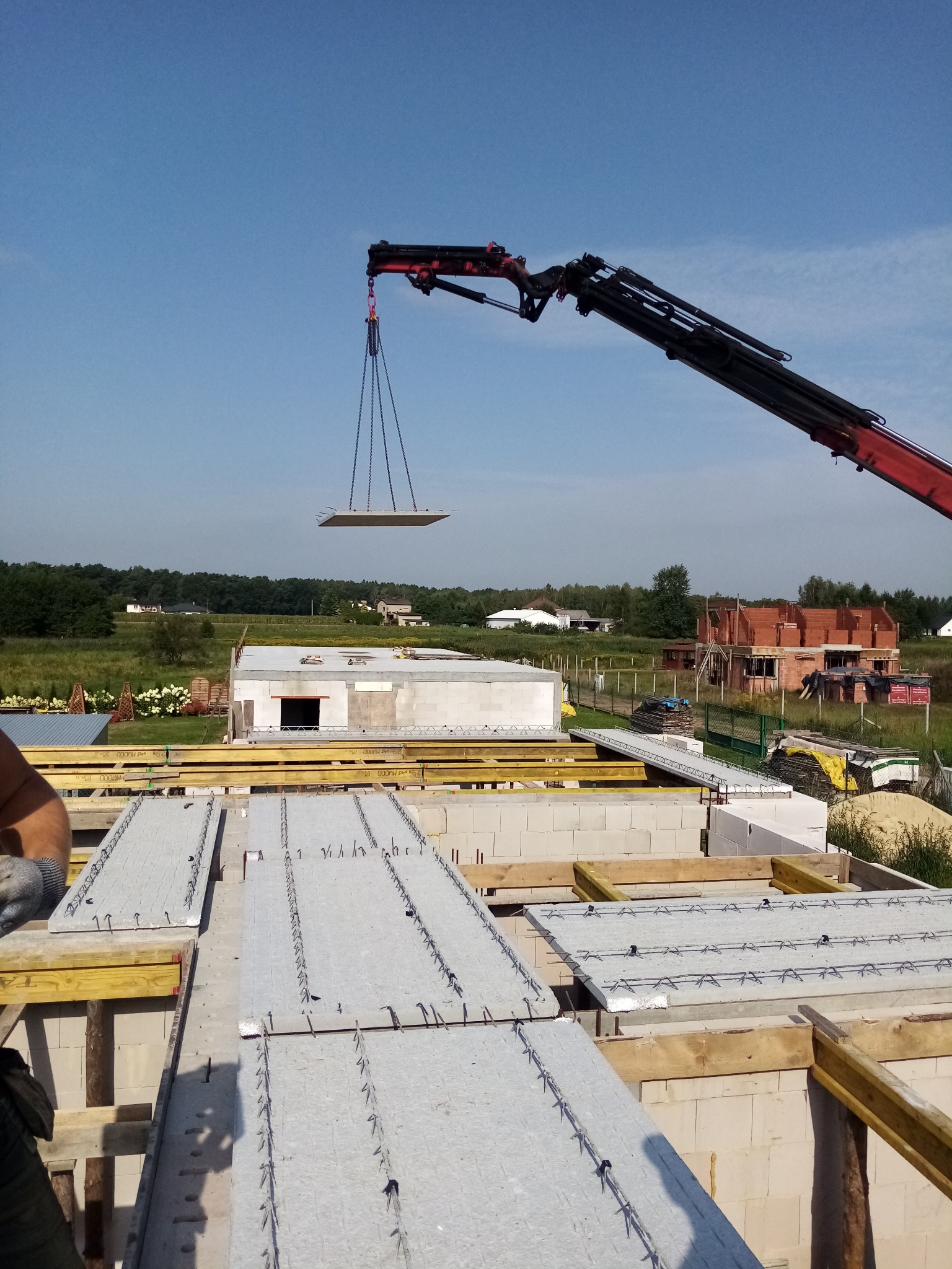 Montaż prefabrykowanych stropów na budowie domu jednorodzinnego przy użyciu dźwigu, w tle widoczny budynek w trakcie wznoszenia ścian z czerwonej cegły.