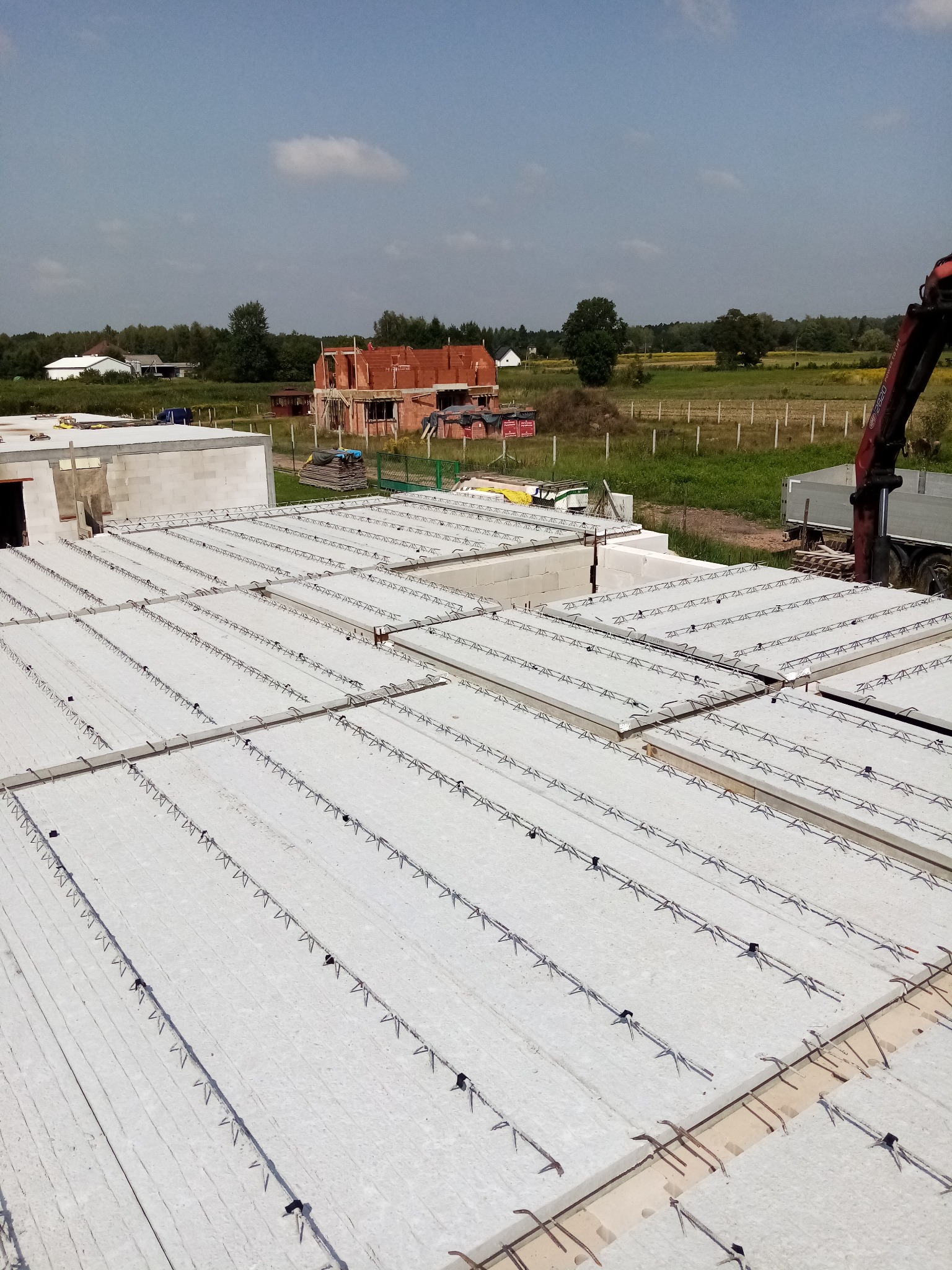 Widok z góry na prefabrykowane płyty stropowe z wystającymi elementami zbrojenia, przygotowane do montażu na budowie domu, z widocznym żurawiem i konstrukcją domu w tle.