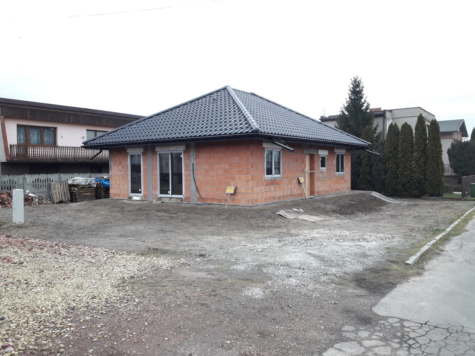 Surowy dom z cegły z ciemnym dachem, widoczne okna i drzwi, teren wokół niezagospodarowany. Budynek w trakcie budowy, otoczony innymi domami i zielenią.