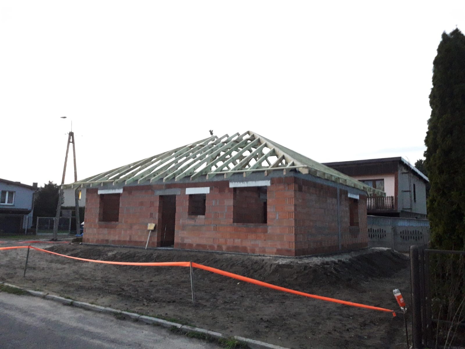 Niedokończony dom z czerwonej cegły z drewnianą konstrukcją dachu na tle nieba. Widoczne zabezpieczenia budowy.