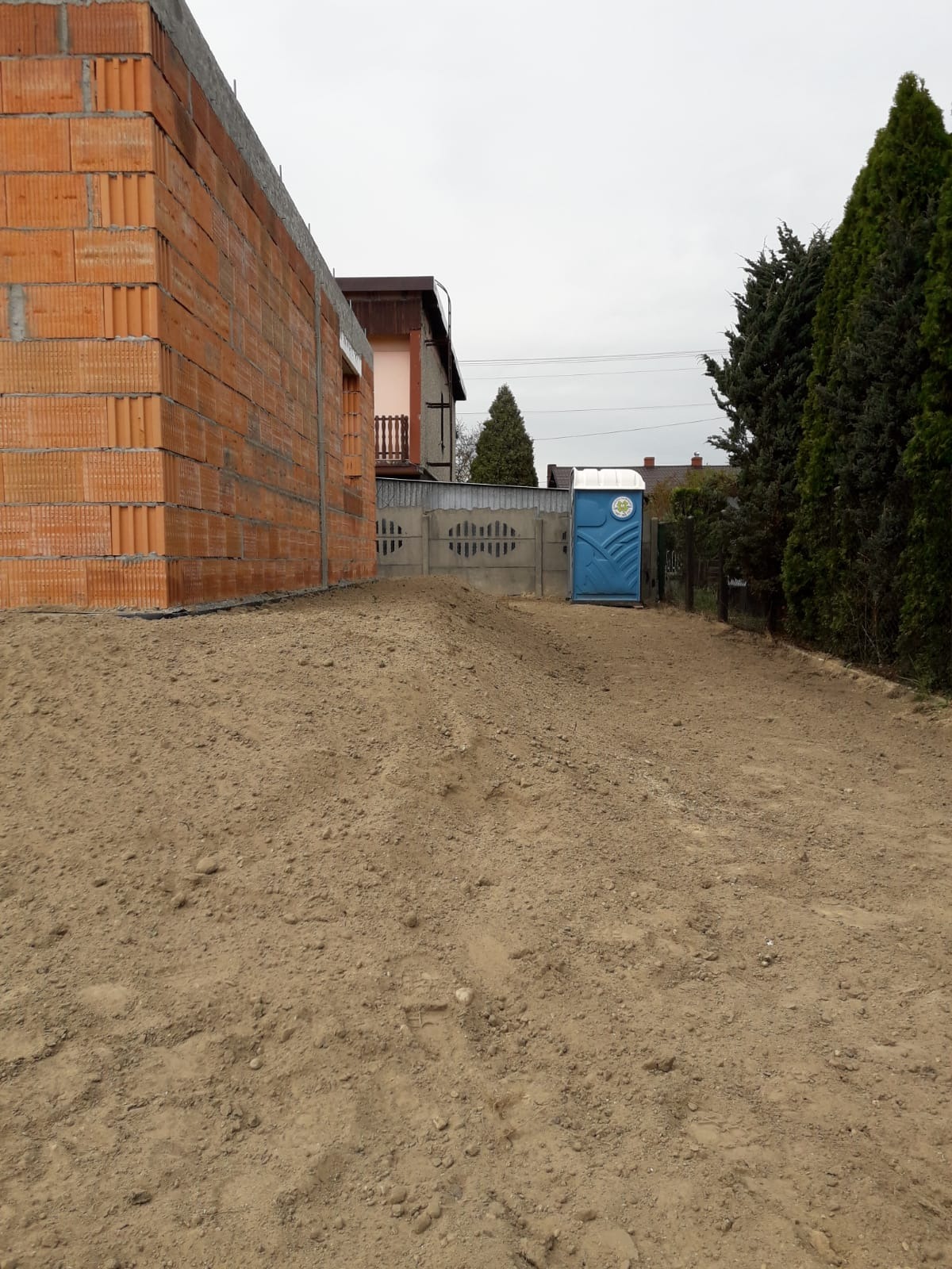 Wznoszona ściana z czerwonej cegły dziurawki, betonowy fundament, niebieska toaleta przenośna i ziemia na placu budowy w pochmurny dzień.