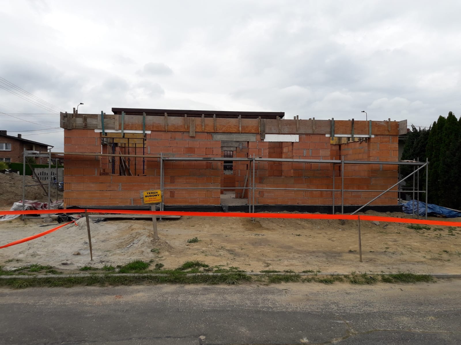 Wznoszenie ścian z czerwonej cegły na budowie domu. Widoczne rusztowania, teren budowy oznaczony taśmą ostrzegawczą i tablica informacyjna. Pochmurne niebo.