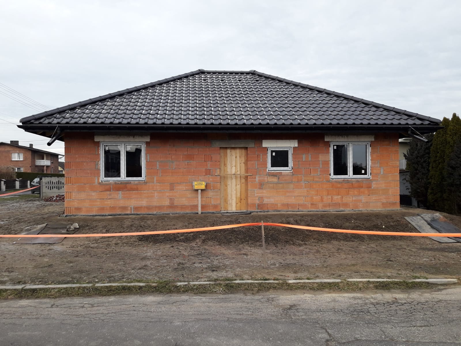 Surowy dom z czerwonej cegły z czarnym dachem, oknami i tymczasowymi drzwiami, otoczony pomarańczową taśmą ostrzegawczą na terenie budowy.