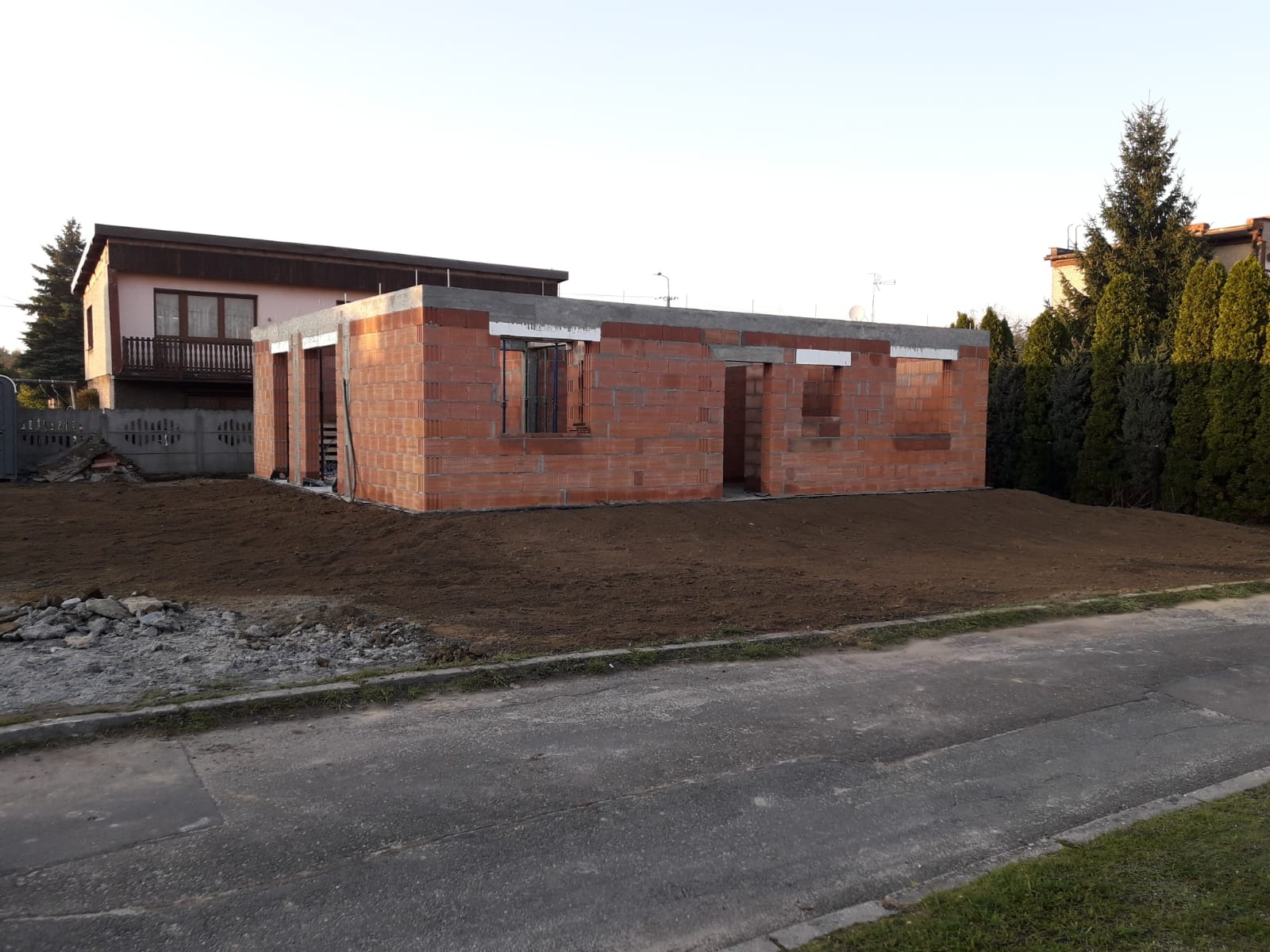 Surowy stan domu z czerwonej cegły, widoczne otwory okienne i drzwiowe, szary fundament, przygotowany teren pod budowę. Dom w trakcie wznoszenia ścian.