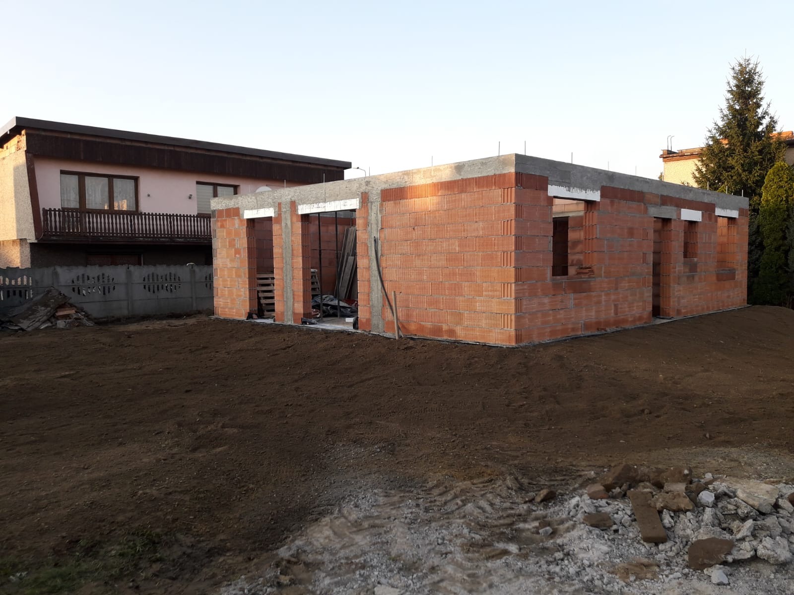 Stan surowy otwarty budynku z czerwonej cegły, widoczne otwory okienne i drzwiowe. W tle dom jednorodzinny i drzewa. Teren wokół budynku wyrównany.