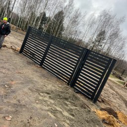 Dawid Sadowski - Czarna, pozioma brama lamelowa w trakcie montażu na błotnistym terenie z widocznym pracownikiem w żółtej czapce i drzewami w tle.