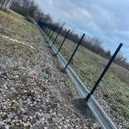 Dawid Sadowski - Długa linia ogrodzenia panelowego z podmurówką betonową na nierównym terenie pokrytym liśćmi, widoczne słupki i siatka, pochmurne niebo w tle.