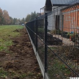 Dawid Sadowski - Nowo postawione czarne ogrodzenie panelowe na betonowej podmurówce oddziela posesję od pola. Świeżo przekopana ziemia wzdłuż ogrodzenia. W tle dom w stanie surowym i pochmurne niebo.