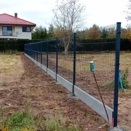 Dawid Sadowski - Nowo postawione, ciemnoszare ogrodzenie panelowe z betonową podmurówką na nierównym terenie. W tle widoczny dom z czerwonym dachem i drzewa w jesiennych barwach. Przy słupku ogrodzenia oparta...