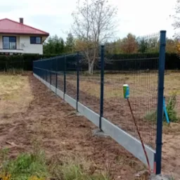 Nowo postawione, ciemnoszare ogrodzenie panelowe z betonową podmurówką na nierównym terenie. W tle widoczny dom z czerwonym dachem i drzewa w jesiennych barwach. Przy słupku ogrodzenia oparta...