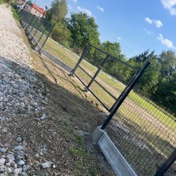 Dawid Sadowski - Fragment nowo postawionego ogrodzenia z siatki, osadzonego na betonowej podmurówce, widoczne z perspektywy żabiej, z kamienistym podłożem po lewej stronie i trawiastym terenem po prawej.