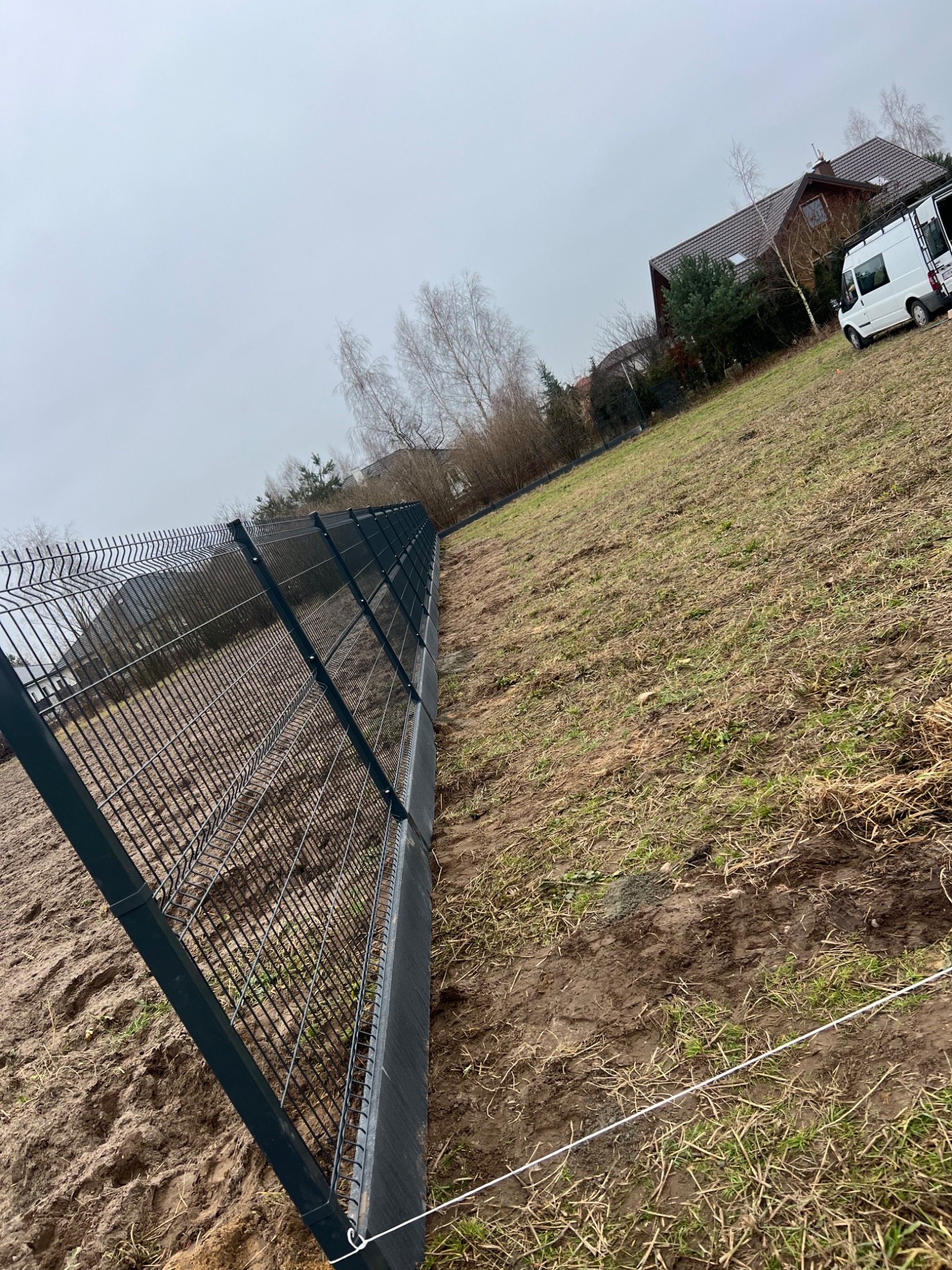 Montaż panelowego ogrodzenia w kolorze grafitowym na nierównym terenie, widoczny biały sznurek wyznaczający linię, dom i biały bus w tle.