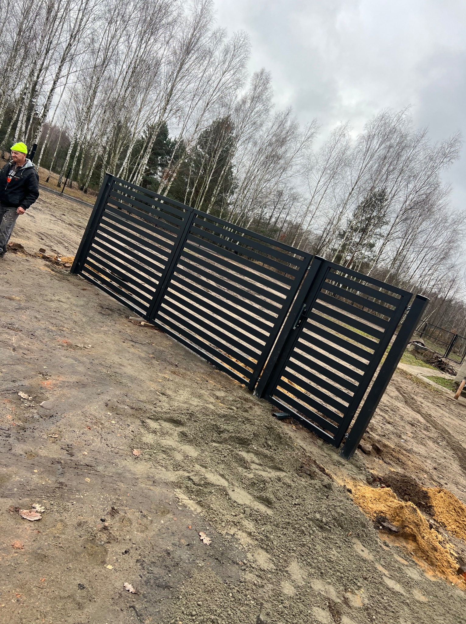 Czarna, pozioma brama lamelowa w trakcie montażu na błotnistym terenie z widocznym pracownikiem w żółtej czapce i drzewami w tle.