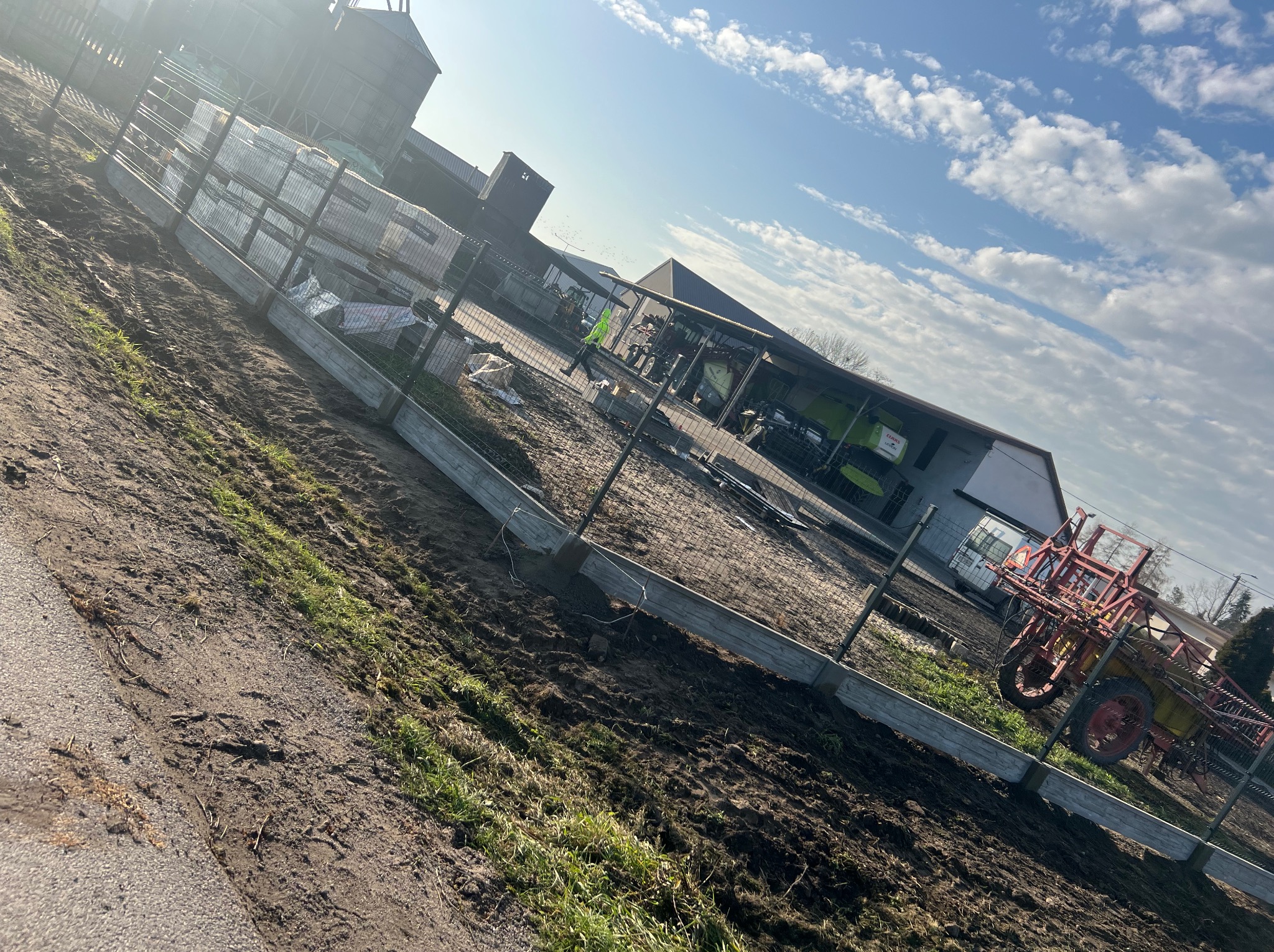 Fragment nowo powstającego ogrodzenia z siatki na betonowych podmurówkach, widoczne role siatki, w tle budynki gospodarcze i sprzęt rolniczy, pochmurne niebo.