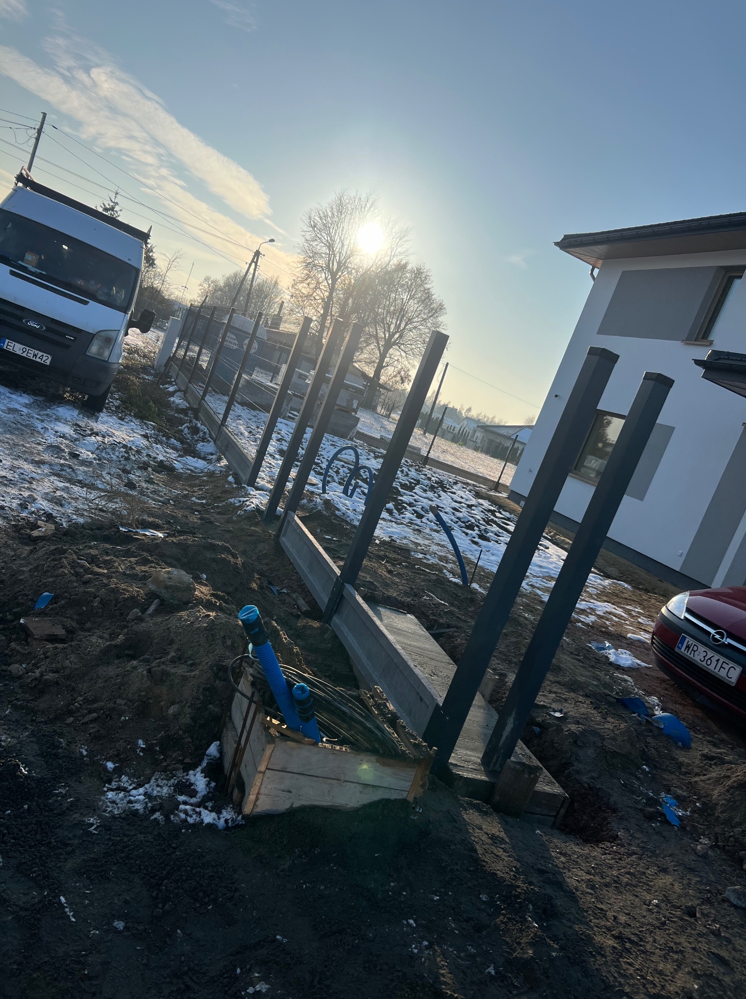 Wykop pod fundament ogrodzenia z widocznymi słupkami i częściowo ułożonymi prefabrykatami betonowymi, na tle samochód dostawczy i dom jednorodzinny w zimowej scenerii.