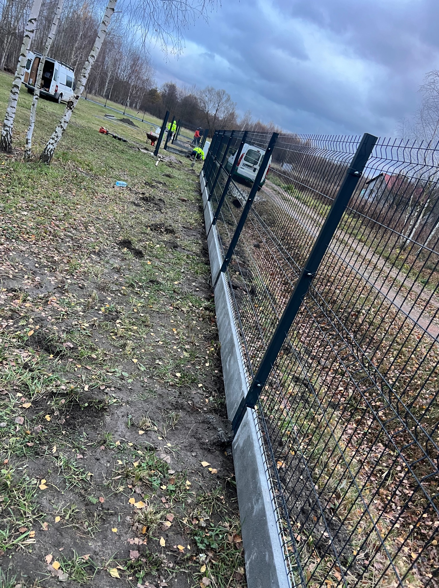 W trakcie instalacji ogrodzenia panelowego na betonowej podmurówce, widoczni pracownicy w odblaskowych kamizelkach, biały bus i brzozy w tle, ziemia pokryta liśćmi.
