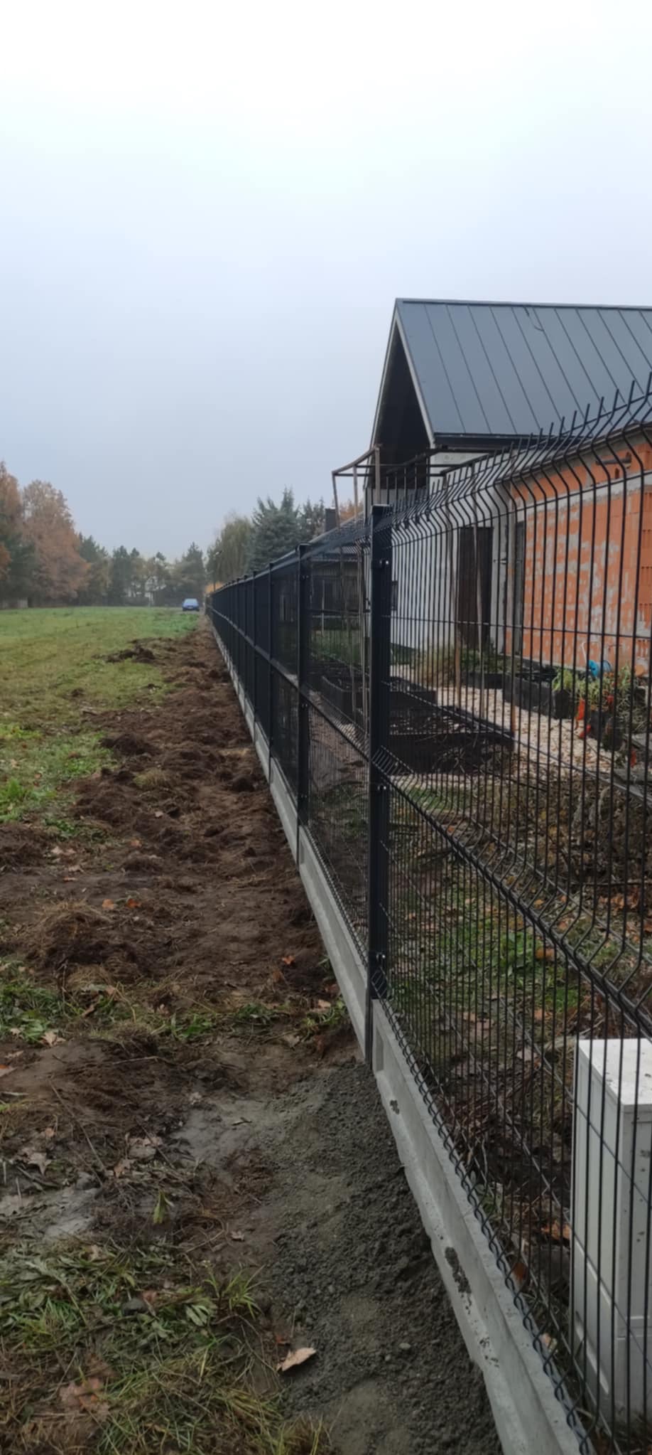 Nowo postawione czarne ogrodzenie panelowe na betonowej podmurówce oddziela posesję od pola. Świeżo przekopana ziemia wzdłuż ogrodzenia. W tle dom w stanie surowym i pochmurne niebo.
