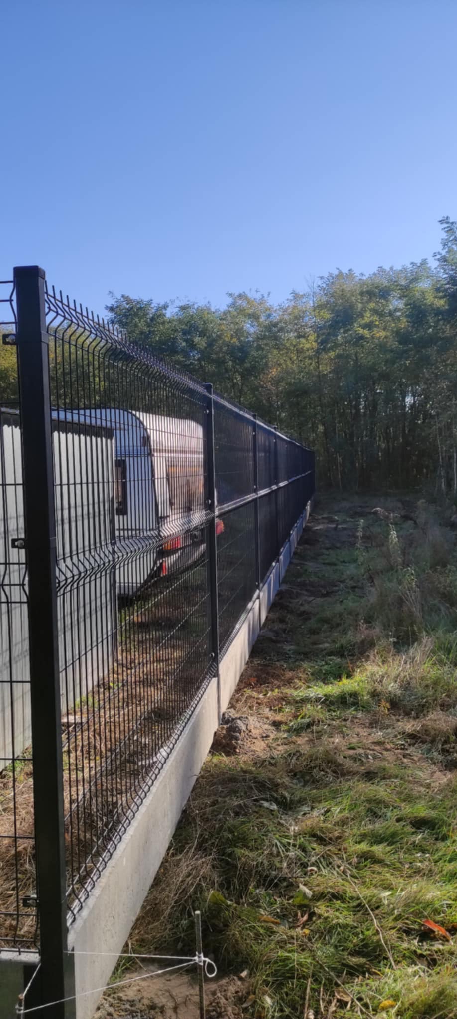 Długi fragment nowoczesnego, czarnego ogrodzenia panelowego z podmurówką betonową, ciągnącego się wzdłuż działki z widocznym zarośniętym terenem i drzewami w tle, w słoneczny dzień.