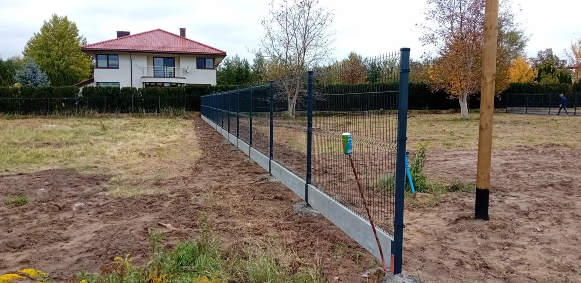 Nowo postawione, ciemnoszare ogrodzenie panelowe z betonową podmurówką na nierównym terenie. W tle widoczny dom z czerwonym dachem i drzewa w jesiennych barwach. Przy słupku ogrodzenia oparta...