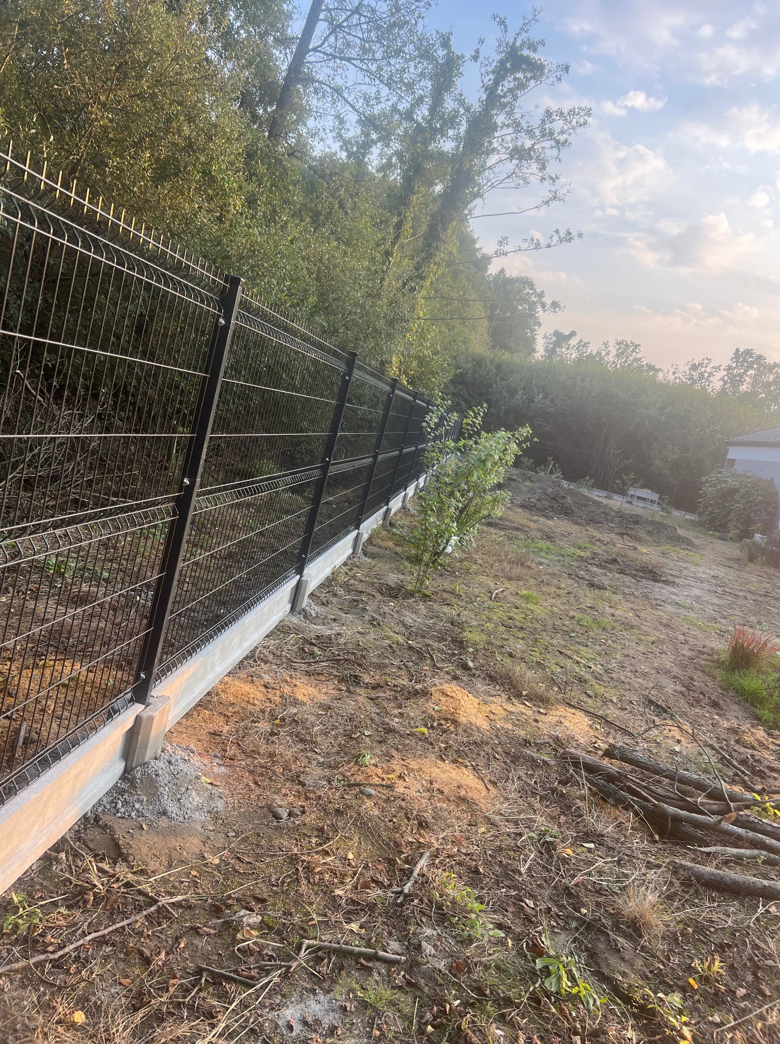 Nowo postawione, czarne ogrodzenie panelowe na betonowej podmurówce, widoczne na tle zieleni drzew i krzewów na nierównym, przygotowanym terenie.