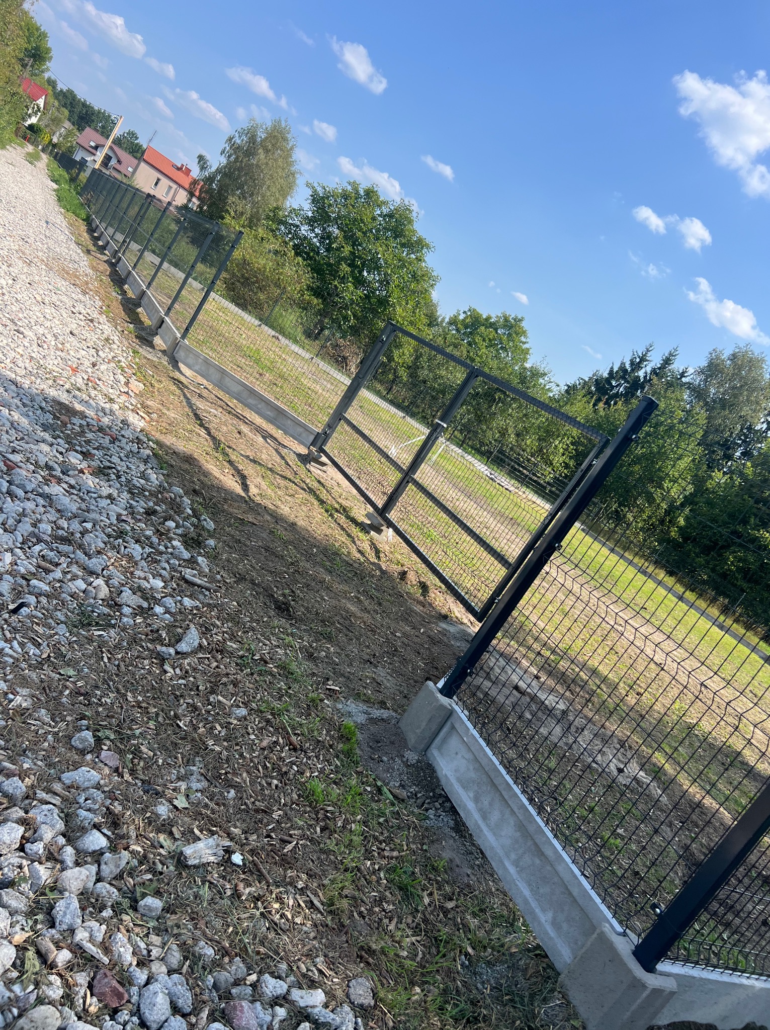 Fragment nowo postawionego ogrodzenia z siatki, osadzonego na betonowej podmurówce, widoczne z perspektywy żabiej, z kamienistym podłożem po lewej stronie i trawiastym terenem po prawej.