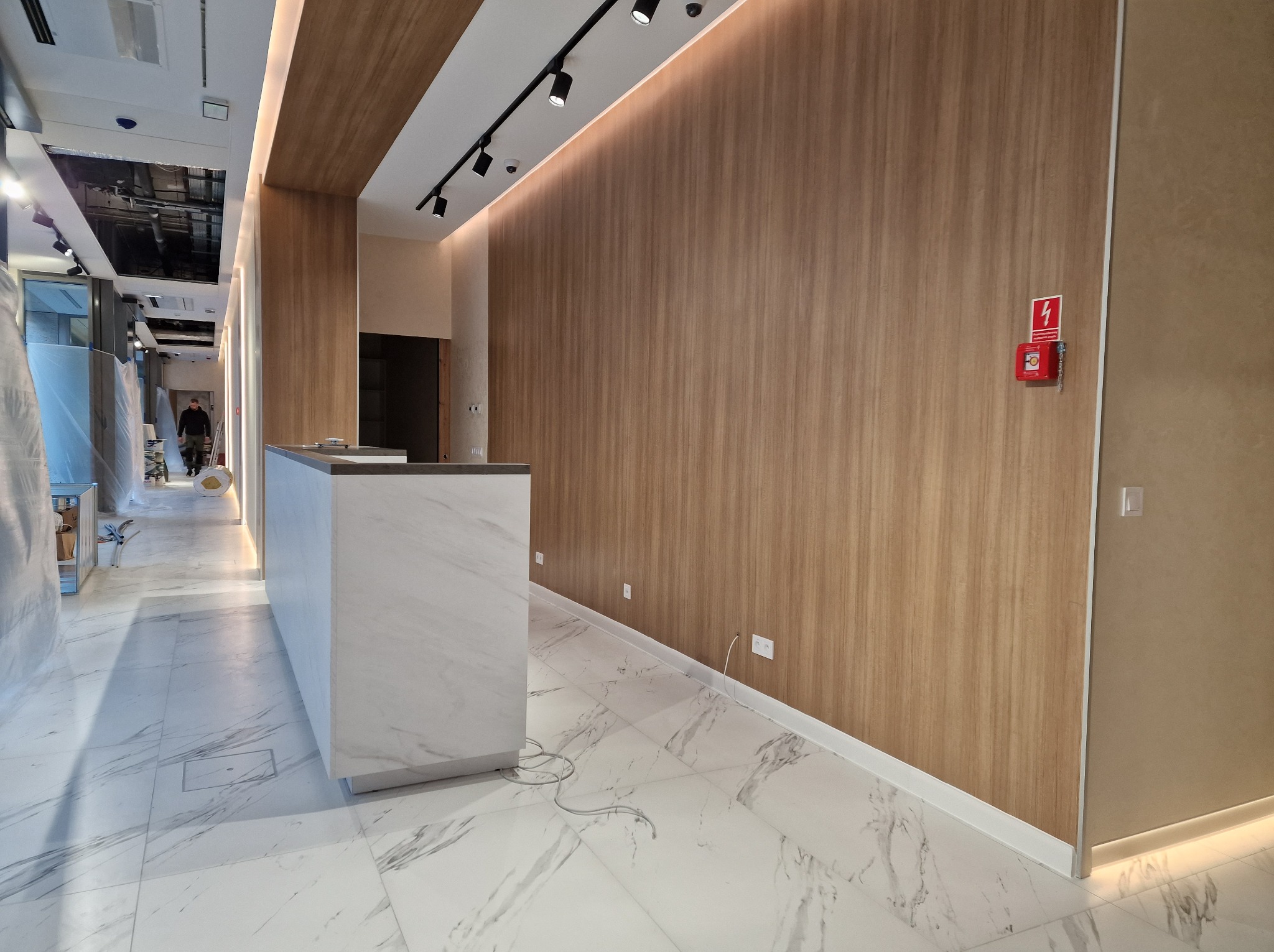 Wnętrze komercyjne w trakcie wykańczania: marmurowa podłoga, drewniana boazeria, biała lada recepcyjna, oświetlenie szynowe i widoczna instalacja wentylacyjna pod sufitem.