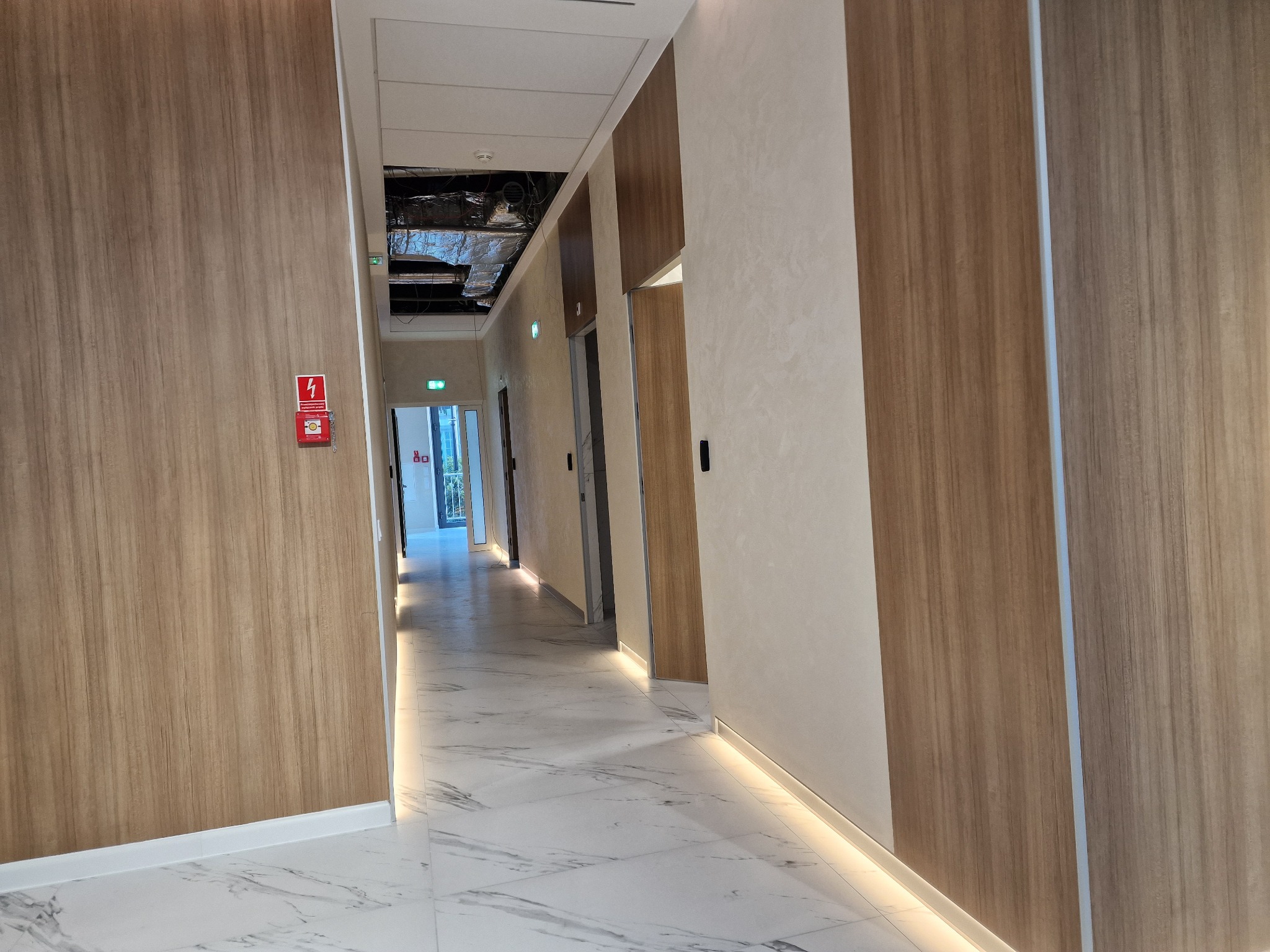 Korytarz biurowy z widocznymi elementami instalacji wentylacyjnej pod sufitem, wykończony panelami drewnopodobnymi i oświetleniem podłogowym.