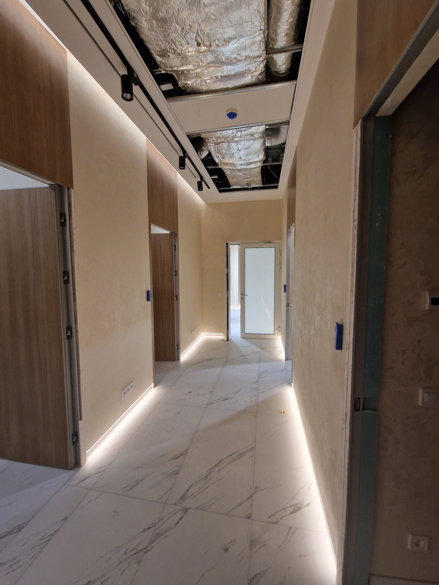 Korytarz biurowy w trakcie wykańczania, widoczna instalacja wentylacyjna pod sufitem, marmurowe płytki podłogowe i listwy przypodłogowe z oświetleniem LED.