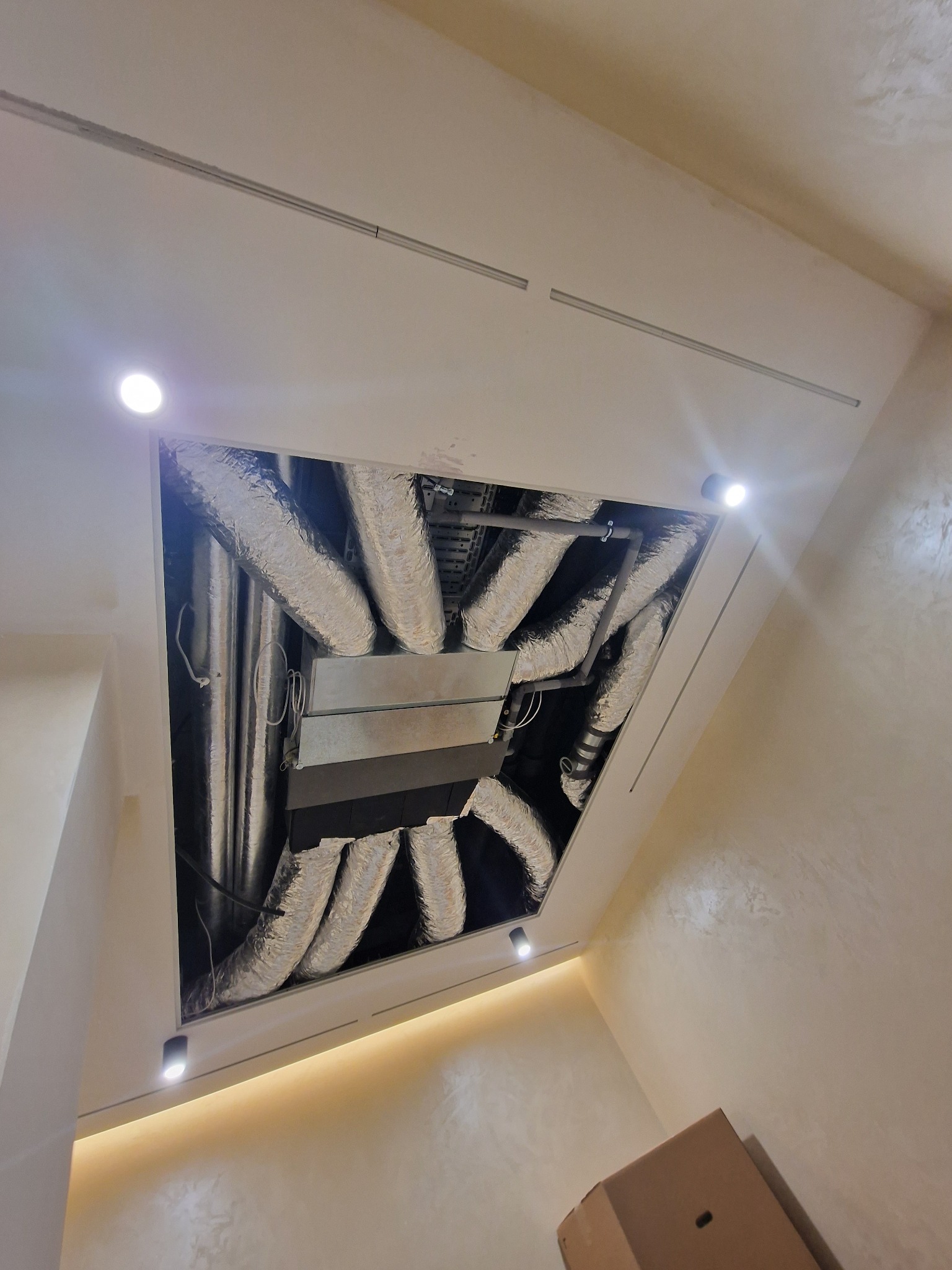 Widok z dołu na otwarty sufit podwieszany z widocznymi srebrnymi przewodami wentylacyjnymi i elementami instalacji, oświetlony punktowymi lampami LED.