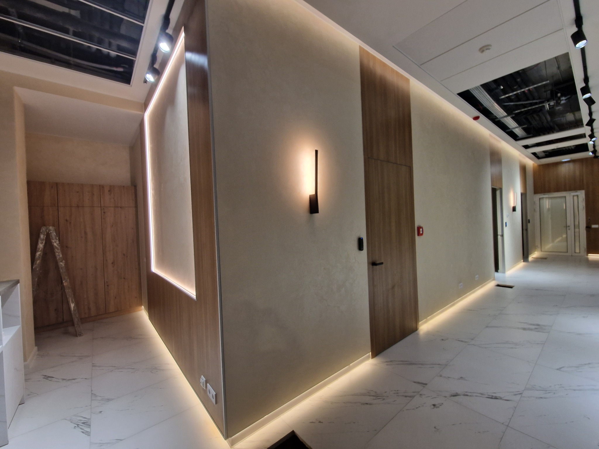 Korytarz biurowy z marmurową podłogą, oświetleniem LED przy podłodze i suficie, oraz minimalistyczną lampą ścienną. Widoczny fragment drabiny oparty o ścianę z szafkami.
