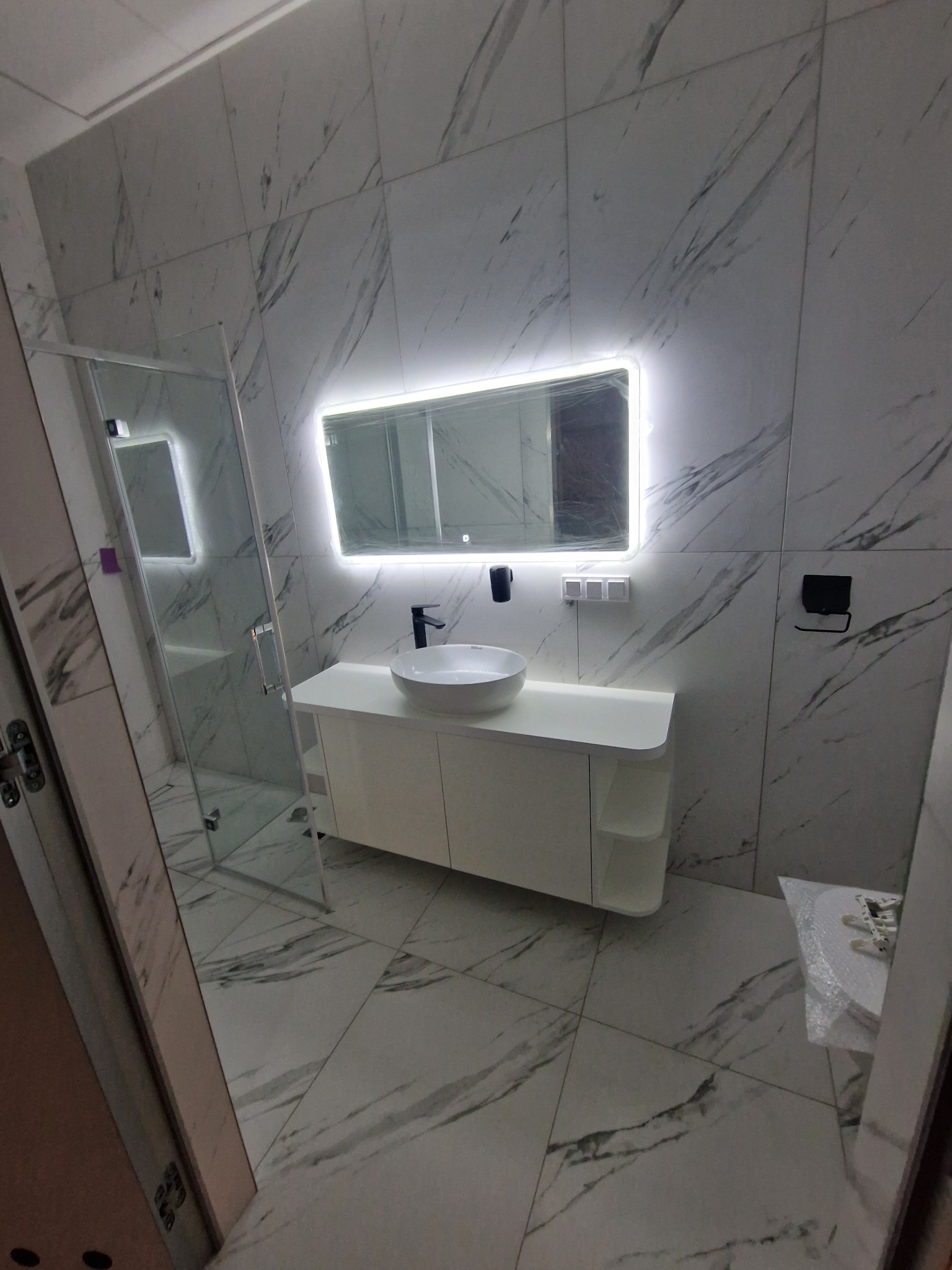 Nowoczesna łazienka z kabiną prysznicową, umywalką nablatową na białej szafce z półkami, podświetlanym lustrem i marmurowymi płytkami.