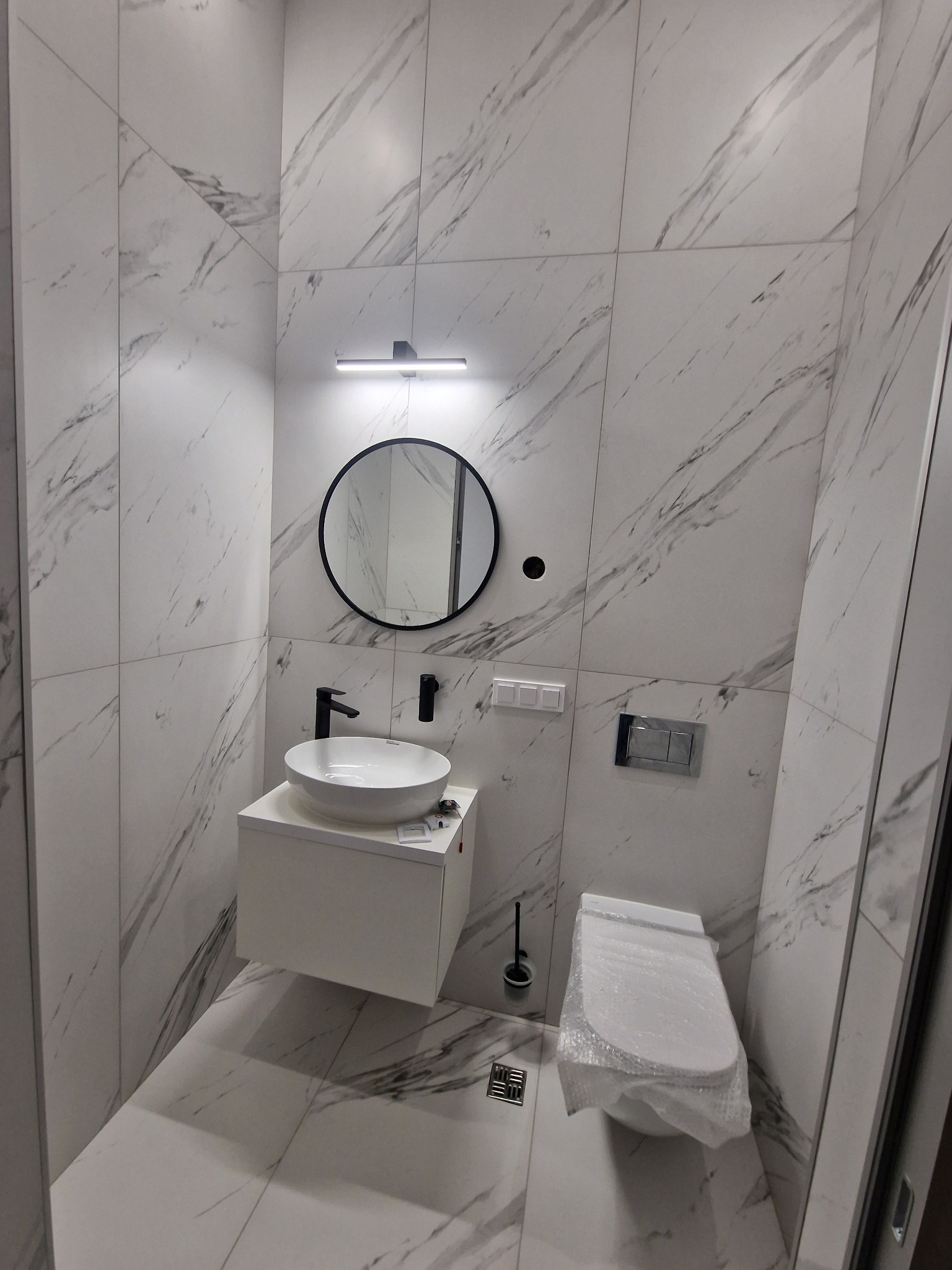 Nowoczesna łazienka wykończona białymi płytkami imitującymi marmur, z okrągłym lustrem podświetlanym lampą LED, białą umywalką nablatową z czarną armaturą i toaletą zabezpieczoną folią ochronną.