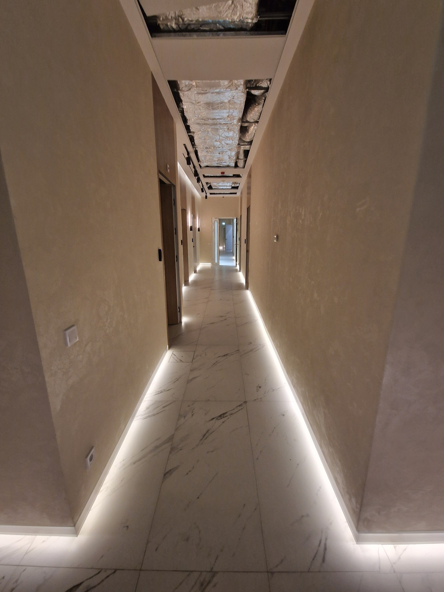 Długi korytarz w budynku komercyjnym z marmurową podłogą, oświetleniem LED wzdłuż ścian i widoczną instalacją wentylacyjną pod sufitem.