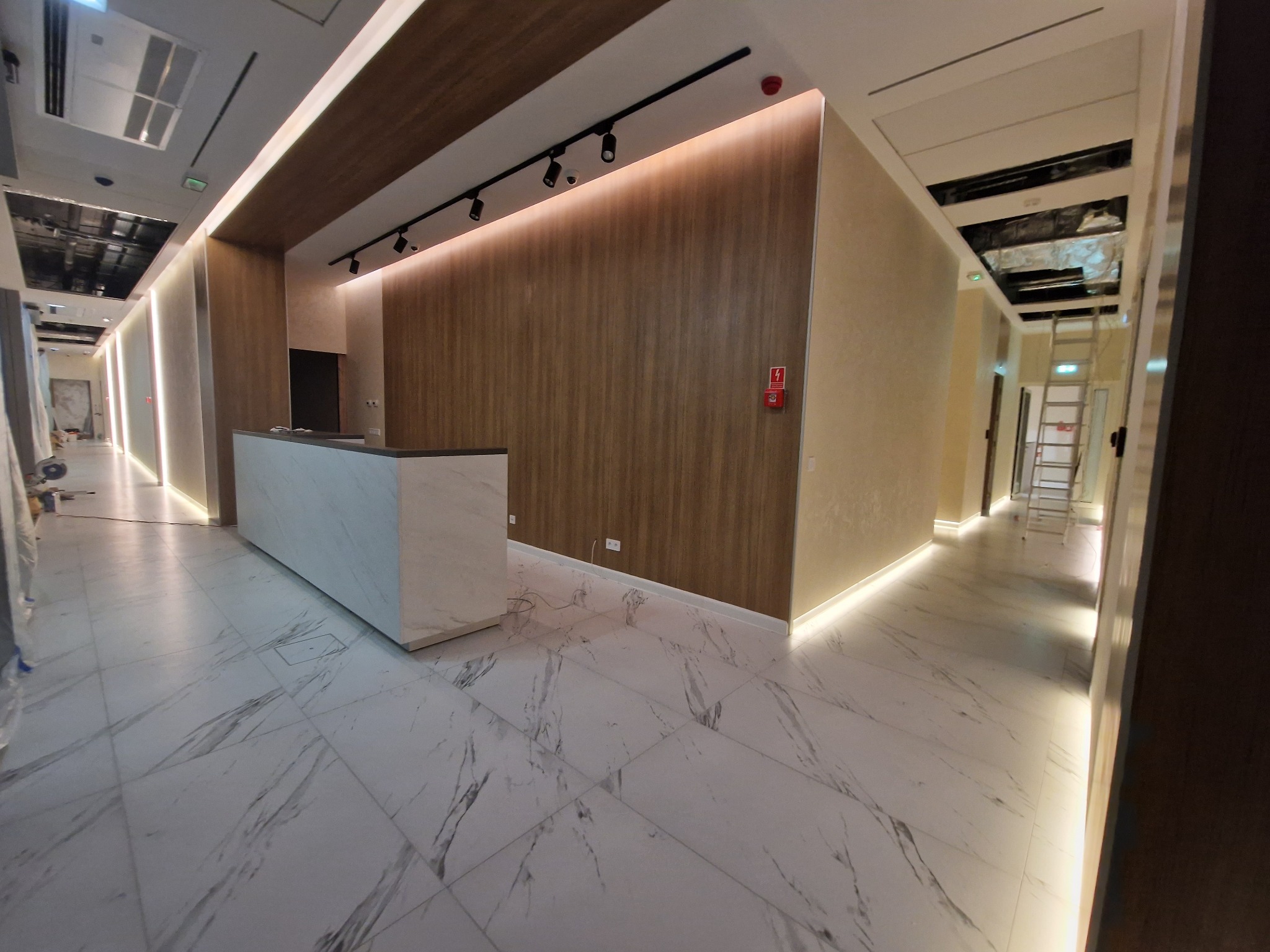 Wnętrze biurowe w trakcie wykańczania: marmurowa posadzka, recepcja z marmurowym blatem, oświetlenie punktowe i liniowe LED, widoczne instalacje pod sufitem w tle.