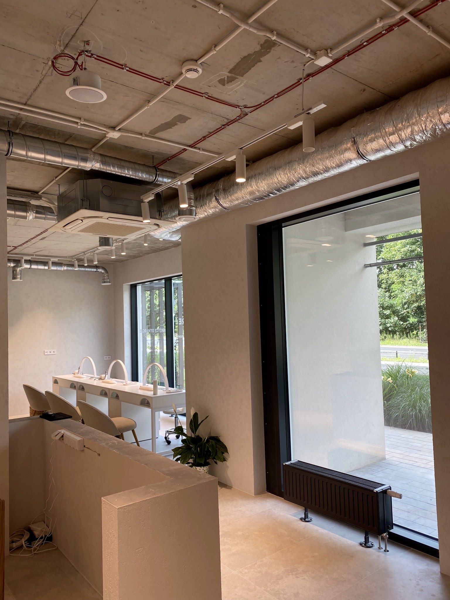 Wnętrze salonu kosmetycznego z widocznymi instalacjami wentylacyjnymi pod sufitem, oświetleniem punktowym, stanowiskami do manicure i grzejnikiem pod oknem.