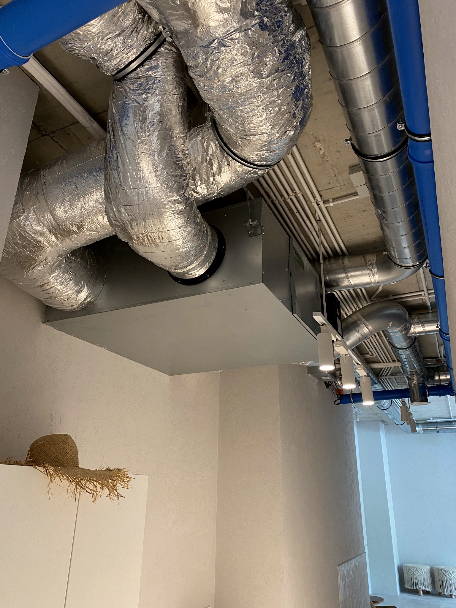 Widok na sufit z systemem wentylacyjnym: srebrne rury izolowane folią aluminiową, niebieskie rury, białe rury instalacyjne, skrzynka wentylacyjna, oświetlenie punktowe, w tle minimalistyczne...