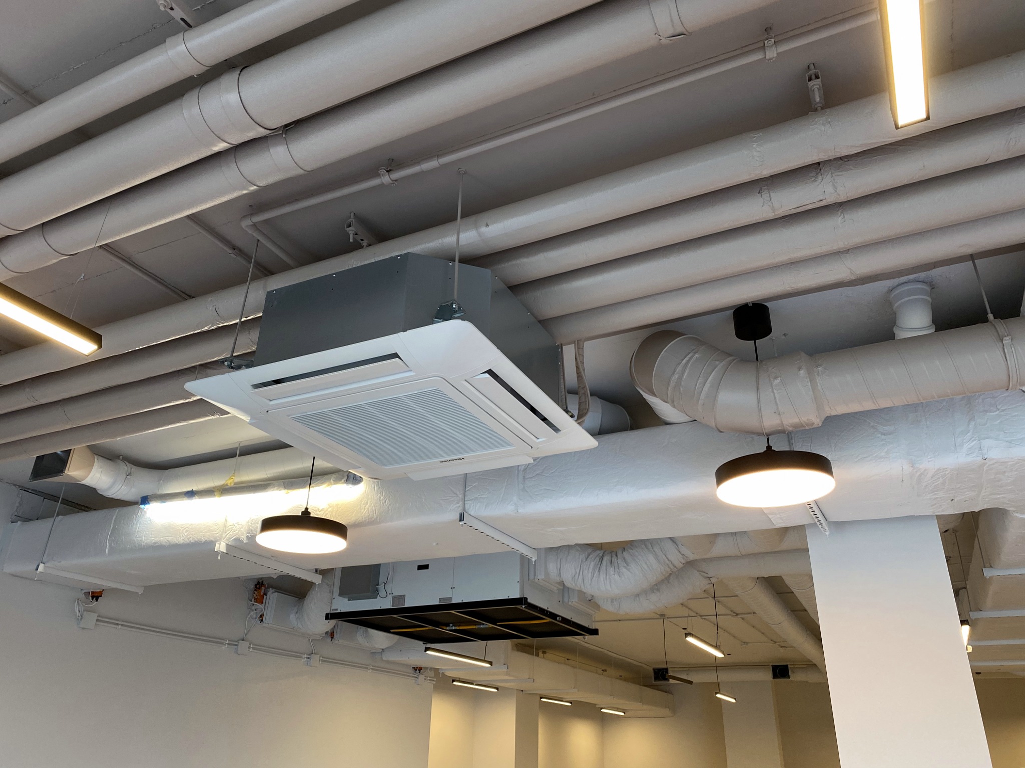 Sufit biurowy z widocznymi instalacjami: rury, kanały wentylacyjne i dwie kasetonowe jednostki klimatyzacyjne z oświetleniem punktowym, pomalowane na biało.