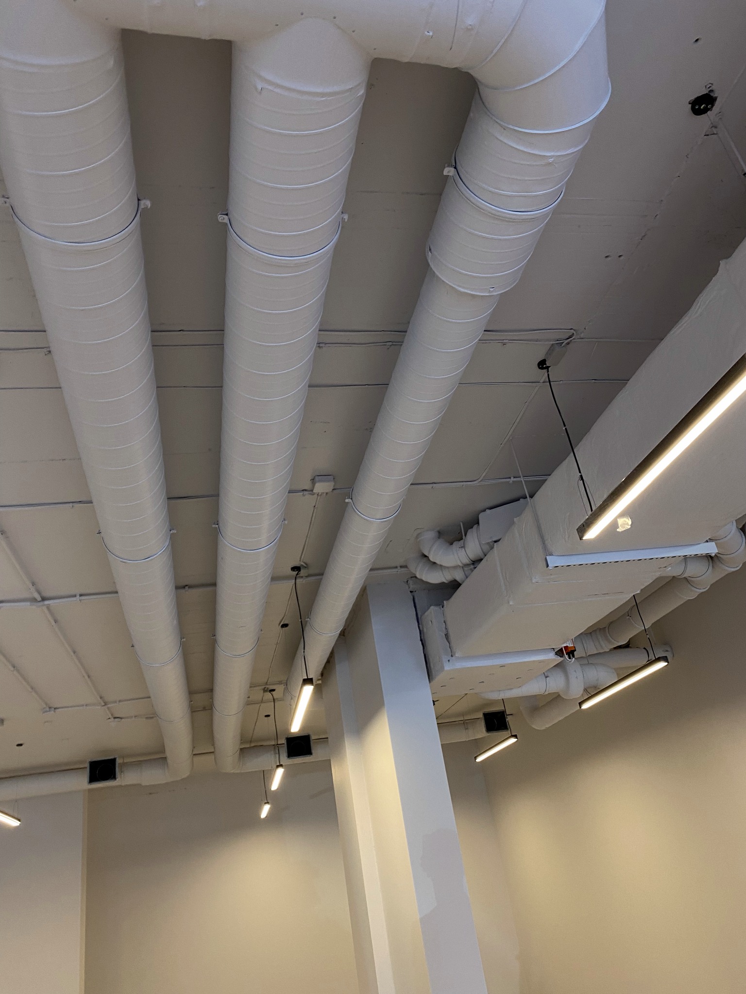 Białe rury wentylacyjne pod sufitem w budynku komercyjnym, widoczne lampy liniowe i mocowania rur.