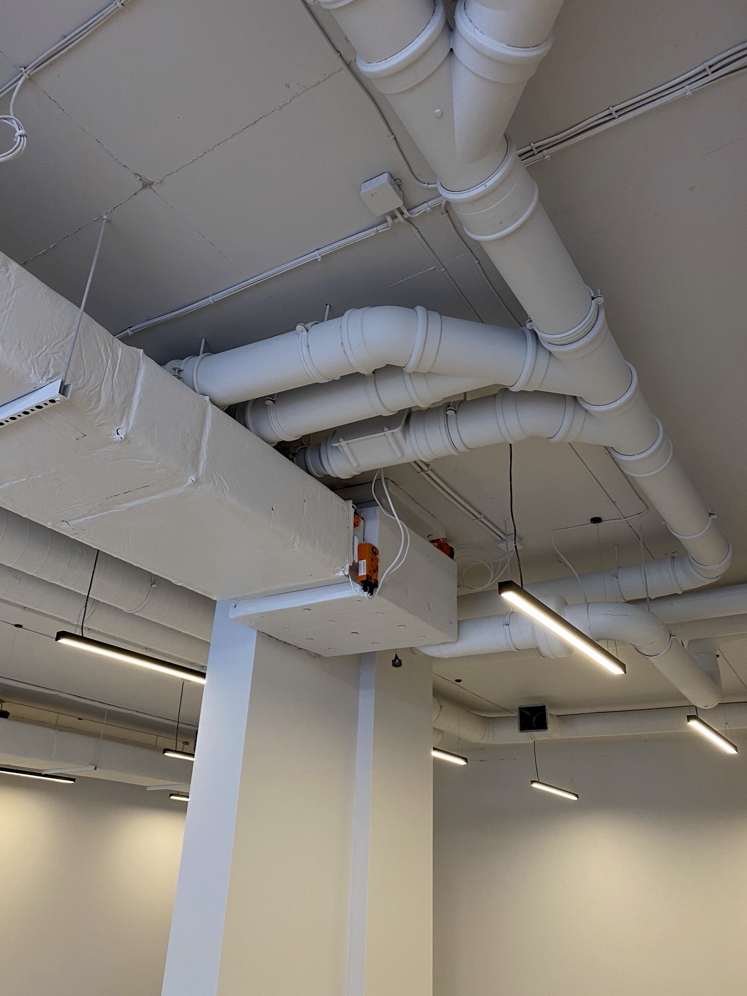 Białe rury wentylacyjne pod sufitem w budynku komercyjnym, widoczne elementy instalacji elektrycznej i oświetlenia liniowego.