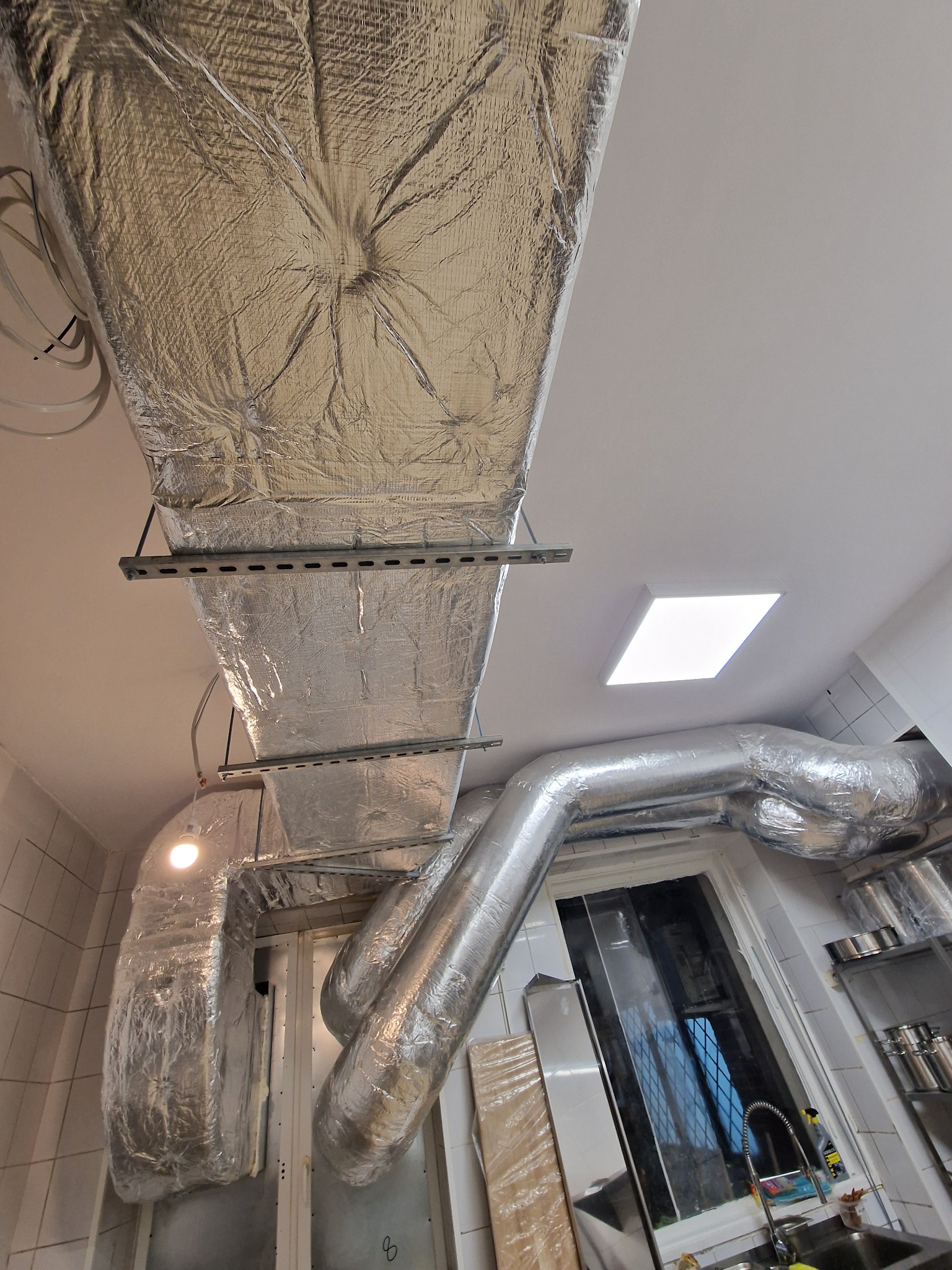 Izolowane kanały wentylacyjne pod sufitem w pomieszczeniu z oknem i zlewem, widoczne metalowe wsporniki i lampa sufitowa.