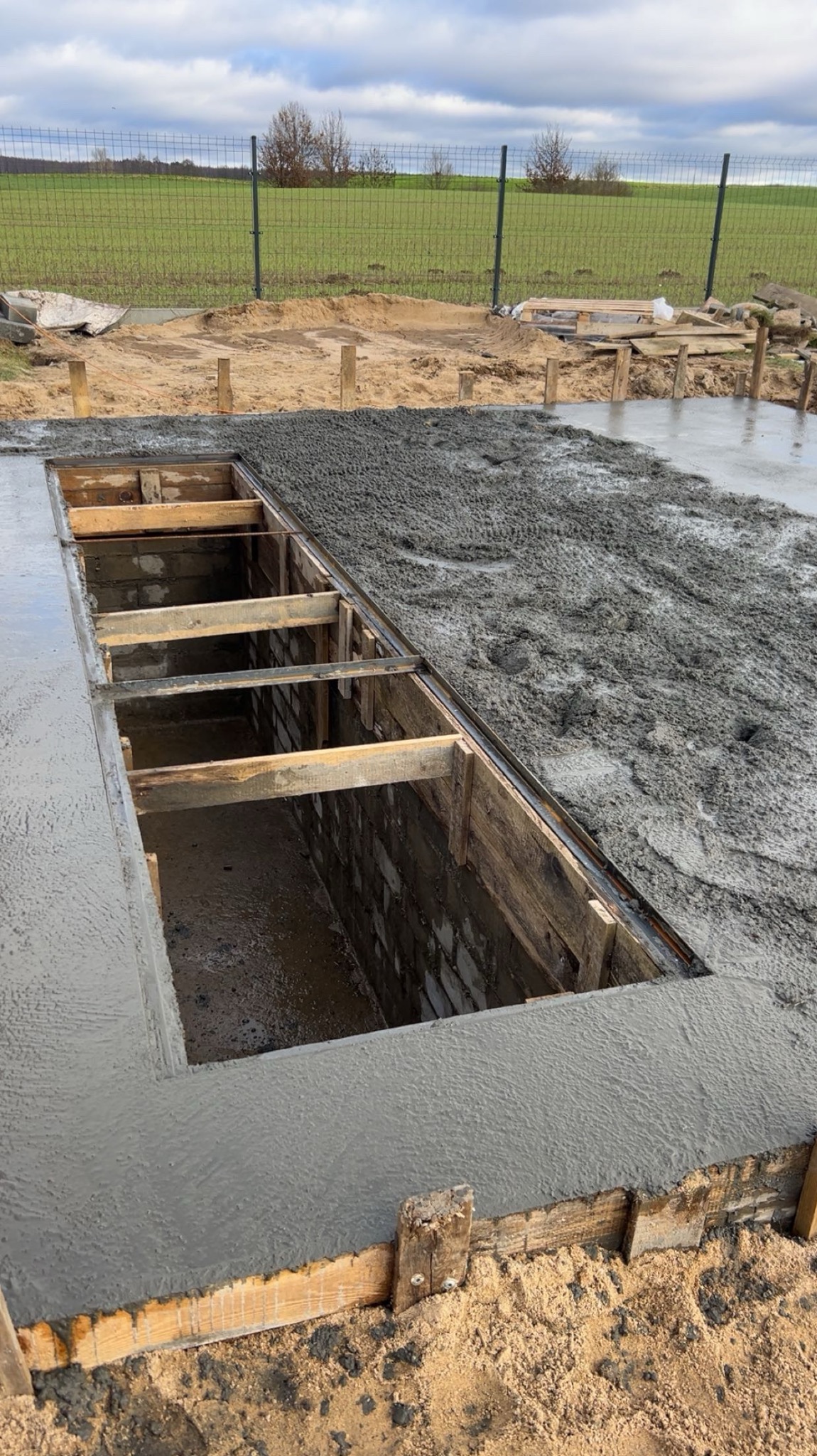 Wylewanie fundamentów z szalunkiem i częściowo rozprowadzonym betonem. W tle widoczne ogrodzenie panelowe i pole.