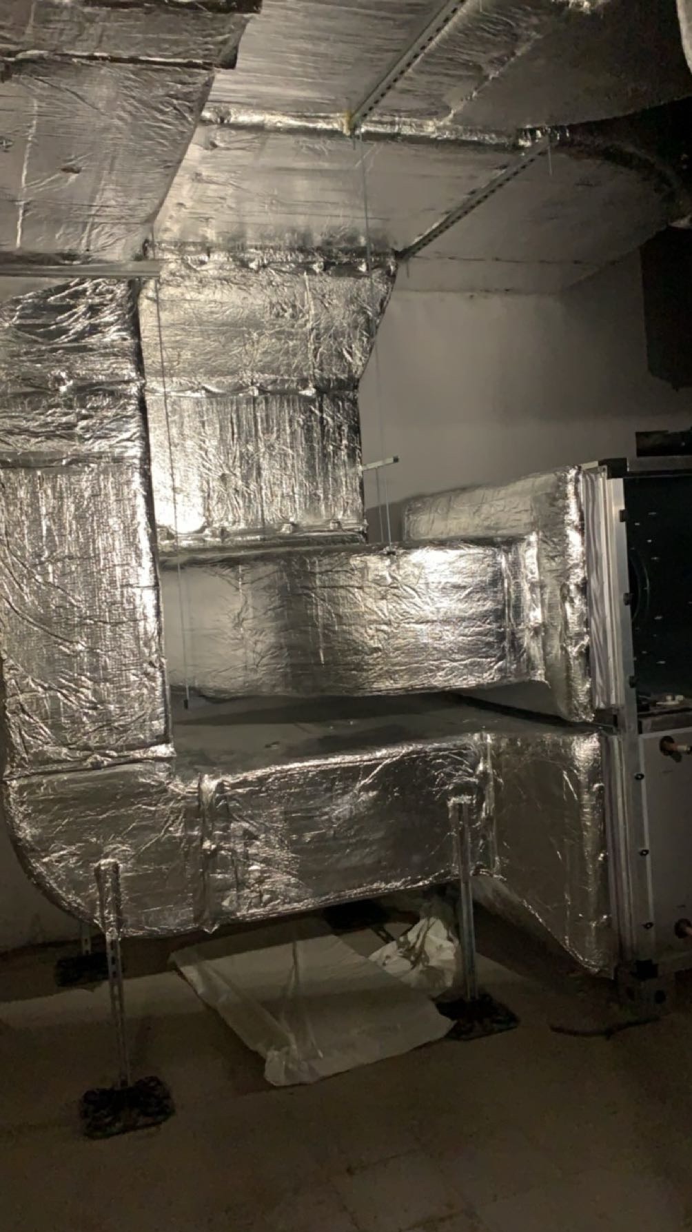 Izolowane kanały wentylacyjne z folii aluminiowej, zamontowane pod sufitem, wsparte na metalowych podporach. Widoczne elementy instalacji i białe worki na podłodze.