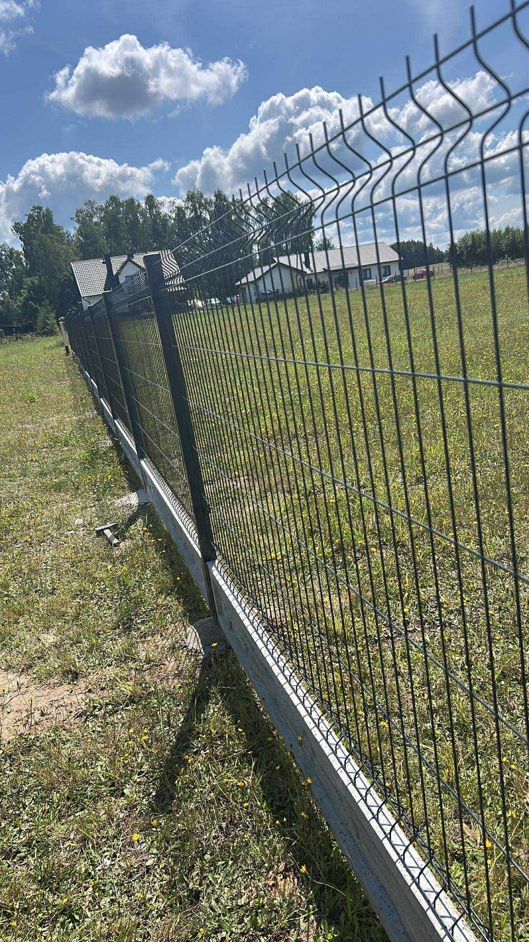 Nowoczesne ogrodzenie panelowe z podmurówką betonową na tle zielonej łąki i domów jednorodzinnych w oddali, widok z boku, słoneczny dzień.