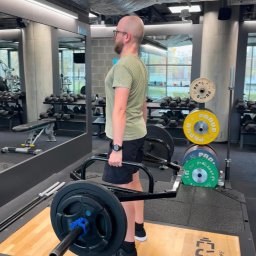 Marcin Stanisławski - Mężczyzna w okularach podnosi ciężar na specjalnym gryfie w siłowni. Widoczne obciążenia, lustro i sprzęt do ćwiczeń. Trening siłowy.