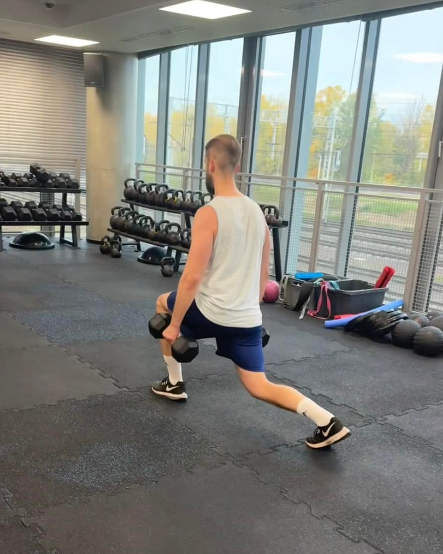 Mężczyzna w koszulce na ramiączkach wykonuje wypad z hantlami w siłowni. W tle okna z widokiem na tory kolejowe i półki z obciążeniami.